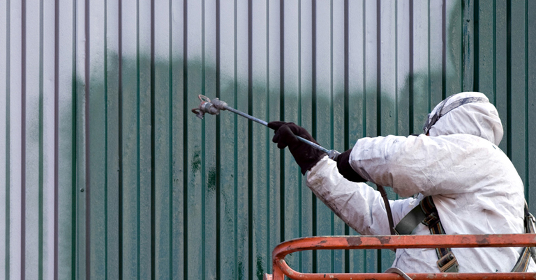 A worker in protective gear spray paints a metal wall green.