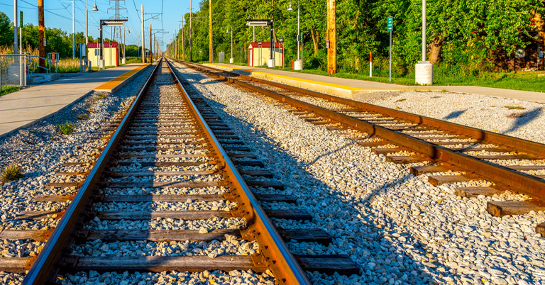 Railroad tracks leading to a small train station surrounded by green trees in bright sunlight.